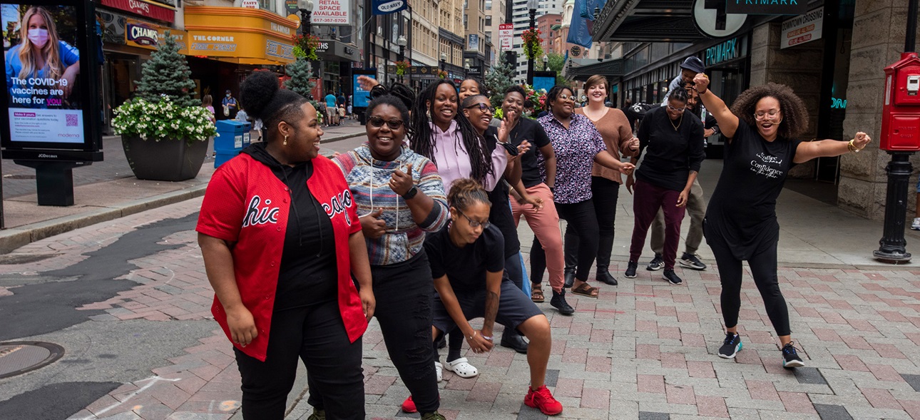 A group of people stand in a curved line on the sidewalk outside Downtown Crossing in Boston. The photo is a happy candid moment with some people smiling, others dancing, and with their arms up.