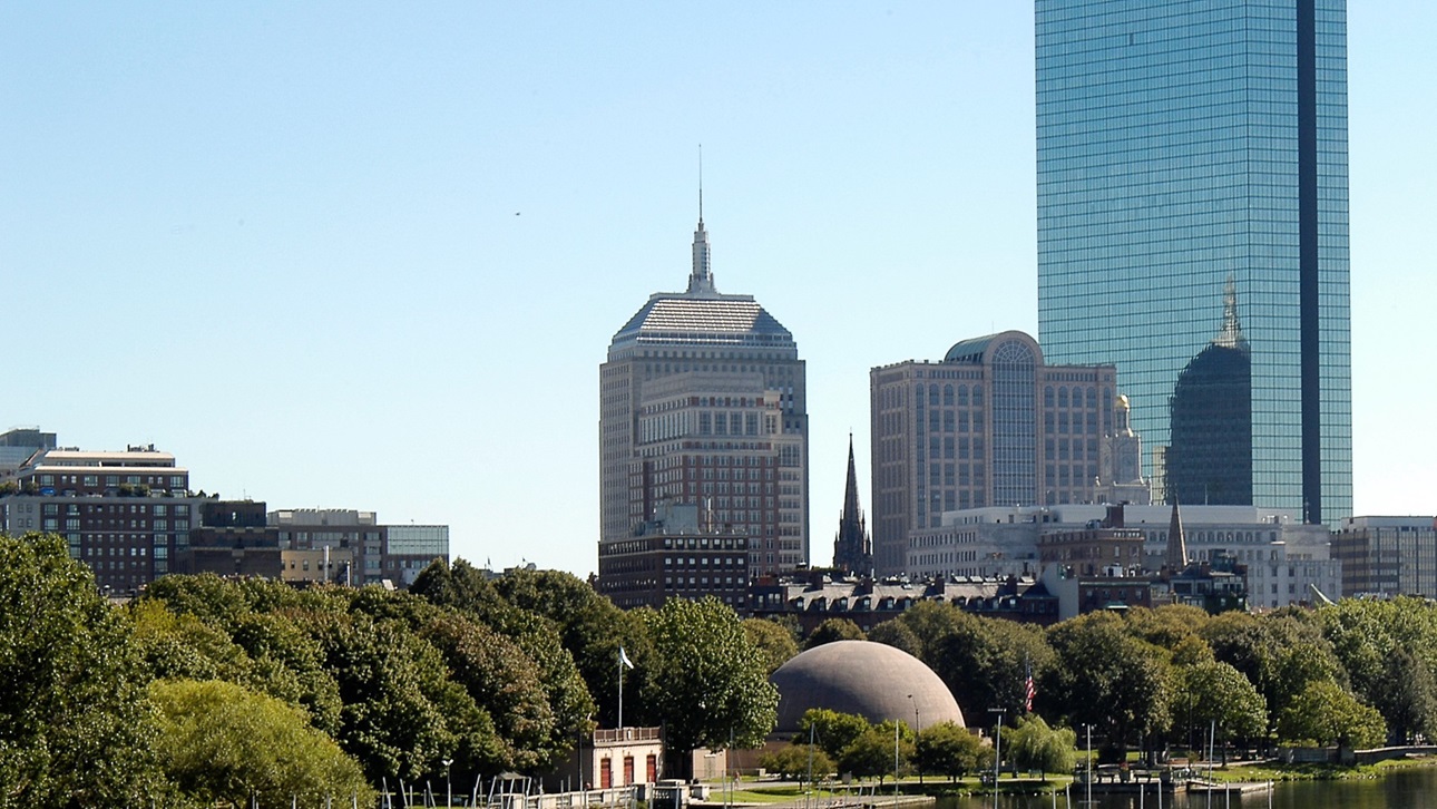 A photo of Boston overlooking the esplanade with many trees in the foreground and a few large buildings in the background