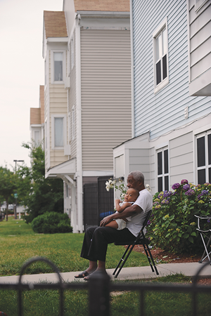 An elderly black man sits on a folding chair on the front walkway to a blue house, he has a toddler in his lap.