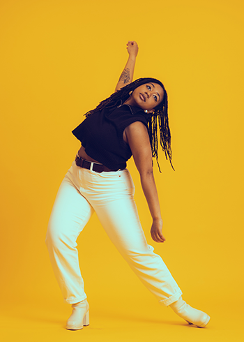 Victoria Lynn Awkward dances in front of a bright yellow background. Her feet are spread apart and her torso is bent so she is leaning to one side with her arms extended vertically