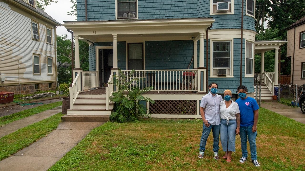 From left to right, Mike, Taheera and Christian Massey are standing in their yard, front of and to the right their teal house in Dorchester. Mike and Taheera are wearing white t-shirts and jeans, and Christian is wearing a blue t-shirt and jeans. The grass is green and brown in their yard. To the left is a paved walkway leading up to their brown and white front porch.