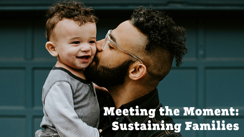 A father, a Latino man, holds his young son and kisses him on the cheek as the child smiles in his father's arms.
